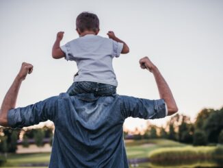 A child sits on an adult's shoulders, both flexing arms playfully. They're outdoors against a sunset sky, evoking joy and bonding in a serene landscape.